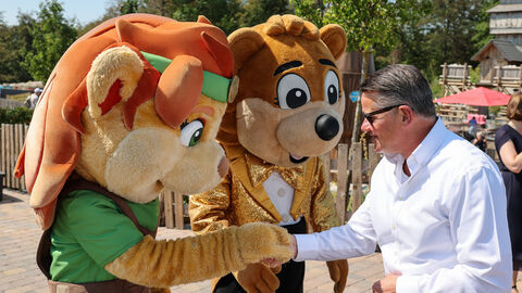 Hessens Ministerpräsident Boris Rhein im weißen Hemd schüttelt einer Bären-Maskottchen-Figur in grünem Gewand die Hand. Im Hintergrund steht ein weiteres Maskottchen im goldenen Glitzer-Frack im Freizeitpark Taunus Wunderland.