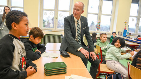 Bildunsminister Schwarz sitzt halb auf einem Tisch und ist Kindern zugewandt, die vor ihren Schulaufgaben sitzen.